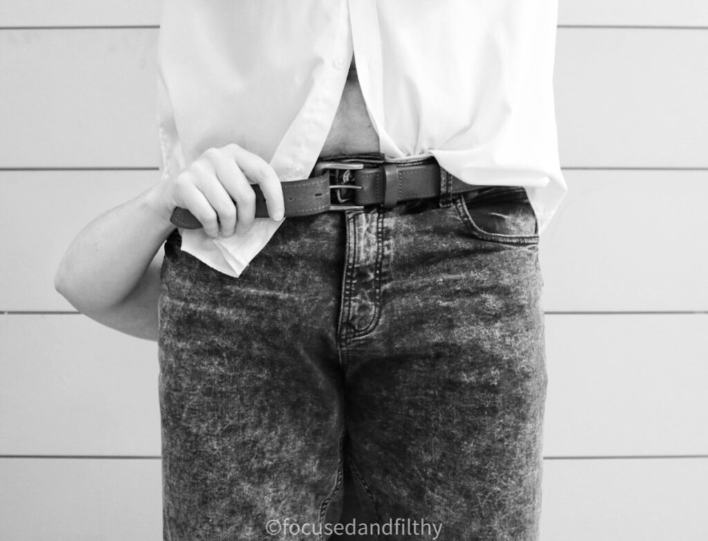 A close up black and white photograph of the middle of a man (from his mid thigh to his mid chest) he’s wearing a white shirt and black jeans and there is a female hand coming from behind him holding on to the end of his belt in the process of undoing it. 