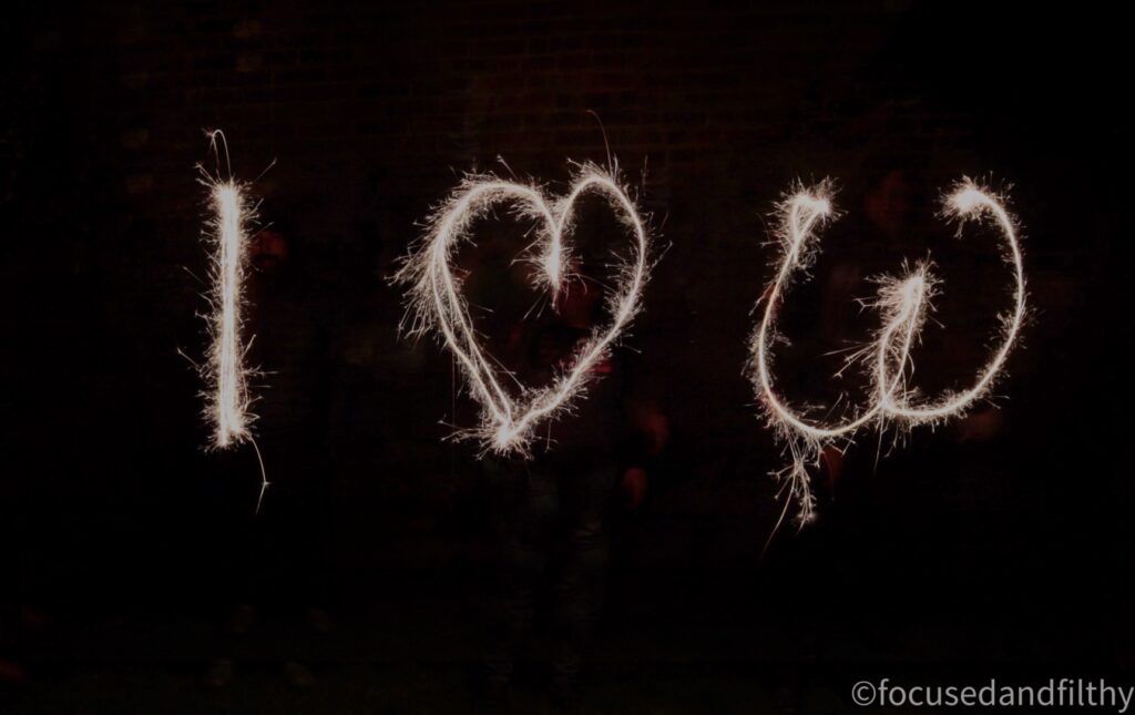 A dark photograph of three shapes made by people with sparklers. From left to right the first one is the capital letter I the second is a heart shape and the third is a slightly badly drawn shape of boobs. 