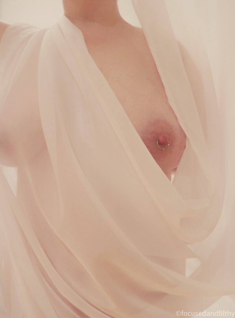 A colour photograph of my naked torso wrapped in a white sheer material. Which just my left nipple sticking out. The image is overall very warm but also very light 