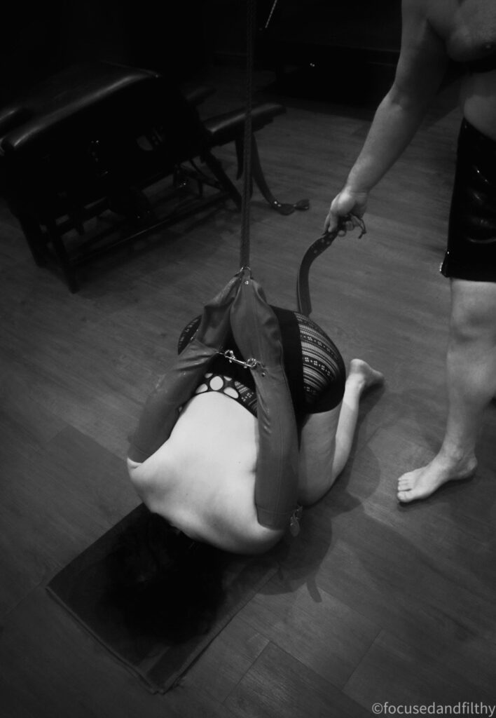 A black and white photograph looking down at a woman kneeling on the floor with her head down and hands tied behind her back attached to a rope which goes up to the ceiling. At the edge of a shot is a man holding a Flogger. 