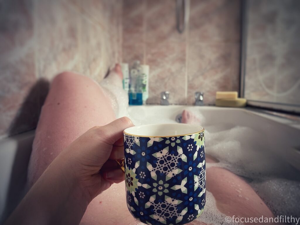 Colour photograph from my point of view. I’m lying in a bath looking down at a mug held in my hands. My left leg is out of the bath on the side. 
