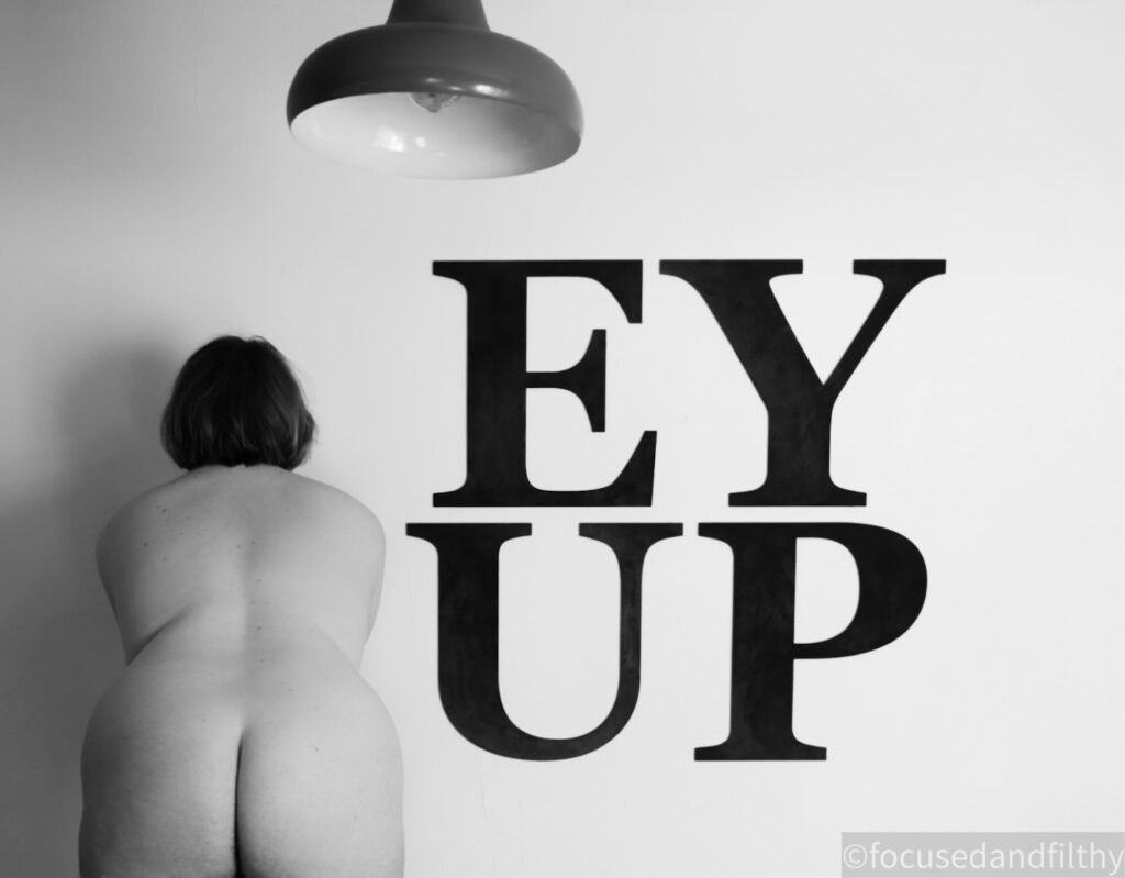 A black and white photograph of a naked woman facing away from you towards a wall. The wall as very large letters on saying EY UP and there is a large industrial style light fitting hanging down too. 