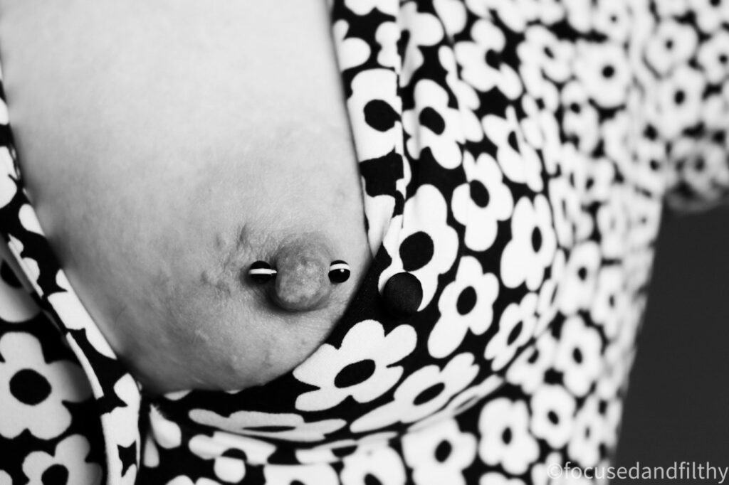 Black and white image of my left side of my chest showing a 60’s flower patterned top with my naked left breast exposed with a black and white striped nipple bar 