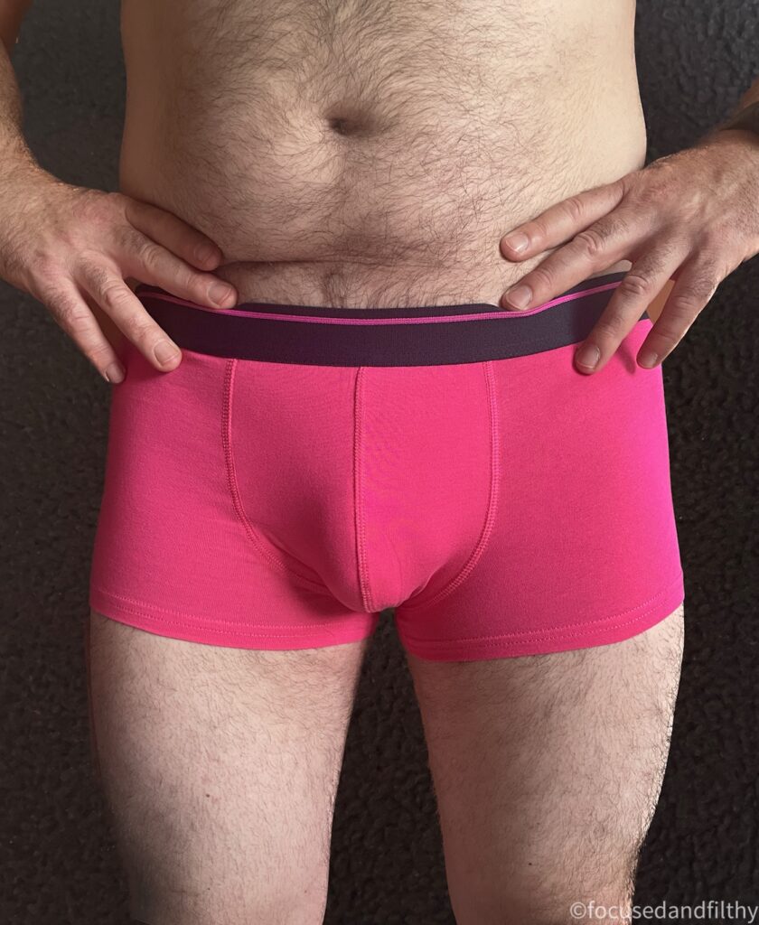 A close up colour photograph of my husbands middle. He’s standing with his hands on his hips and only wearing a pair of bright fuchsia pink trunks (pants) 