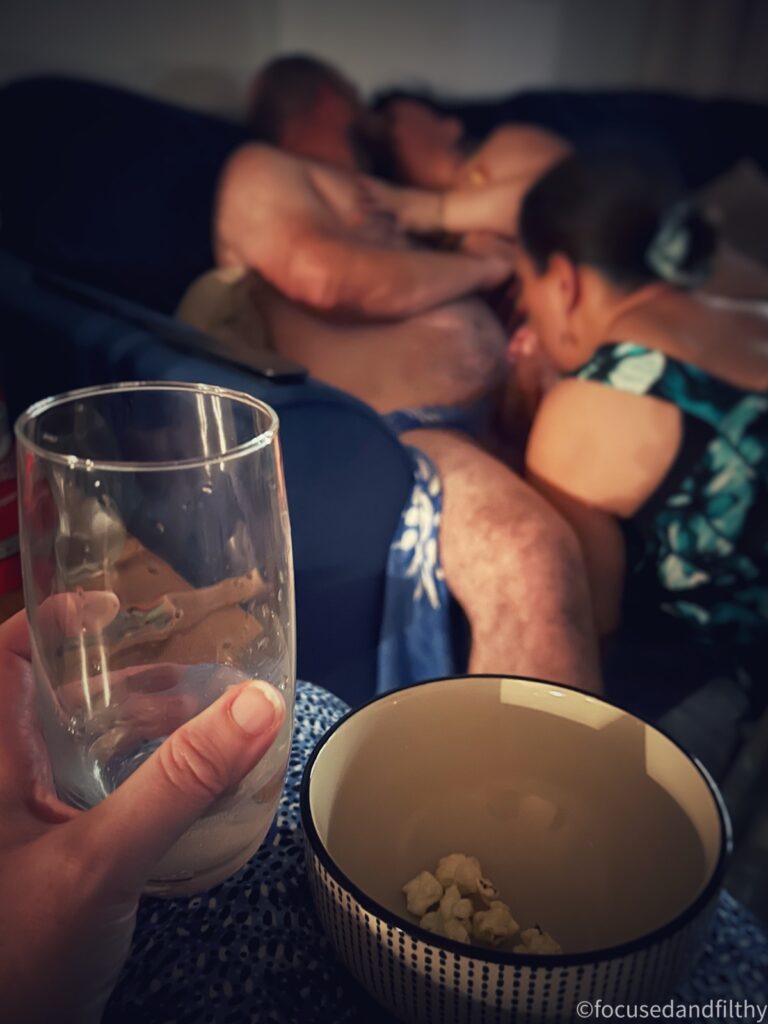 A colour photograph with the foreground in focus on my gin and tonic and a near empty bowl of popcorn and in the blurred background there are three people. One obvious naked male sat down kissing a woman to his left while another one is knelt between his legs sucking his cock 