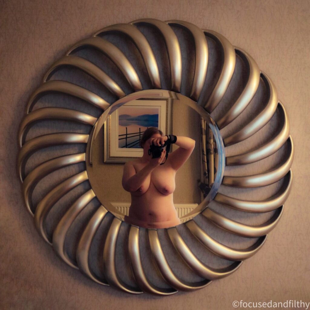 Square colour photograph of a large round ornate mirror. The edges have curved lines like a shell all the way round. In the mirror I’m stood naked holding my large digital camera up to my face to take the image. You can just see my torso. 