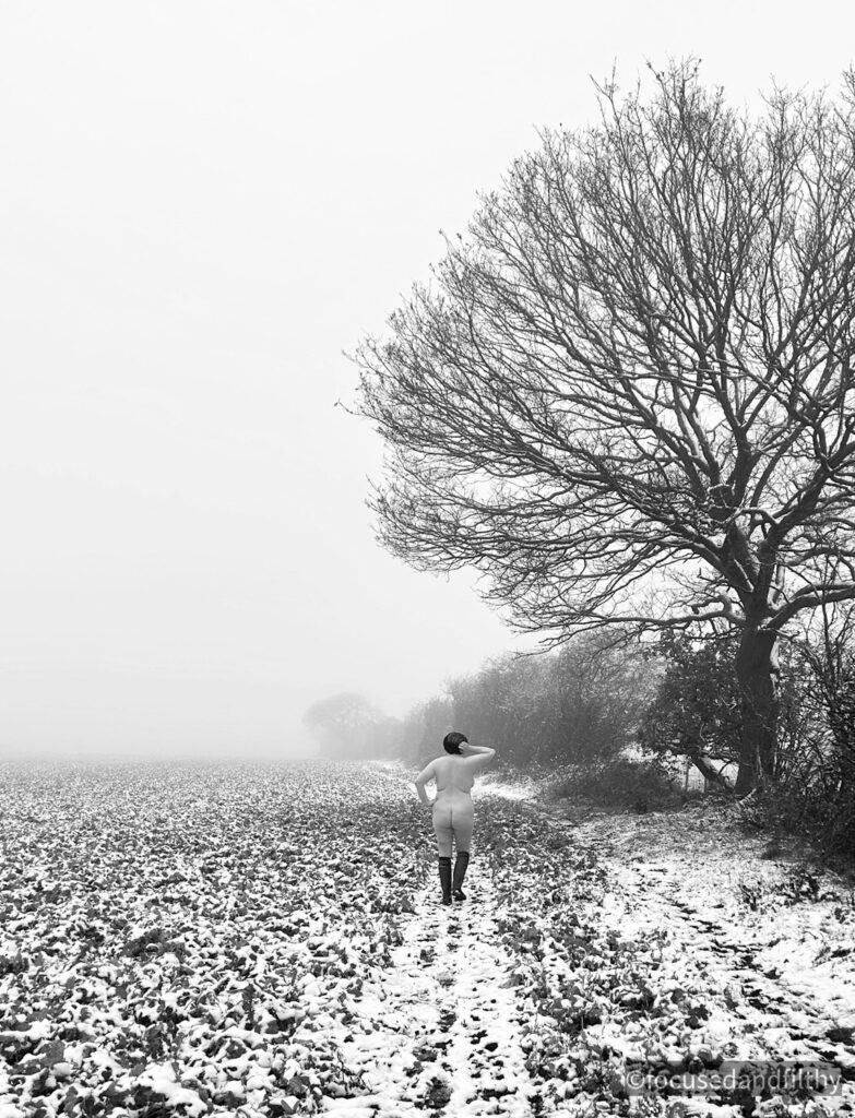 A black and white photograph of a snowy landscape with a path next to a field and tree lined hedge line. On the path is a naked woman walking away. Only wearing wellies and a furry hat. Her pale skin similar colour to that of the snow! 