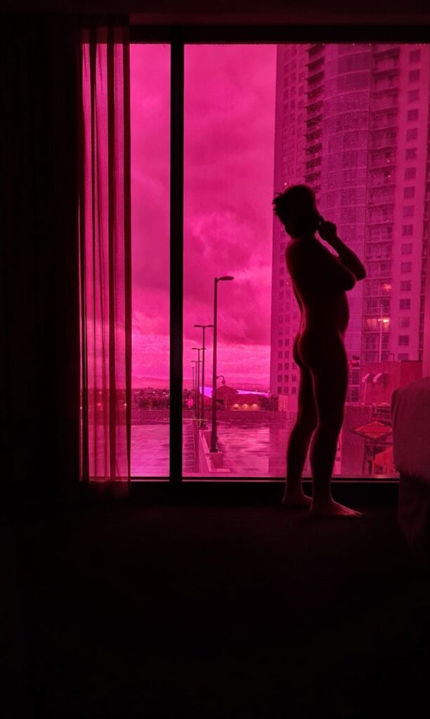 A colour photograph of a very large hotel window with a really magenta colour to it.  Outside it another tall hotel building and in silhouette is a naked figure standing sideways with their hand up to their chin contemplating the view. With a really cute arse. 