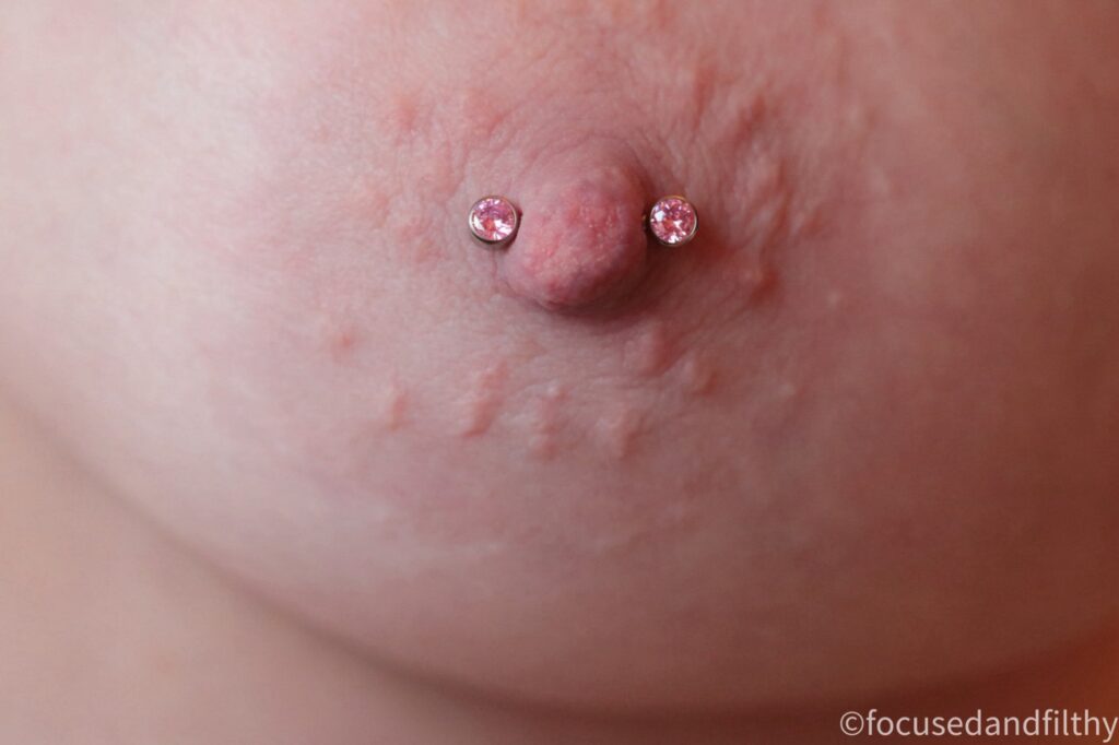 Close up colour photograph of my left breast and nipple showing a nipple bar with pink sparkly stones in each side. 