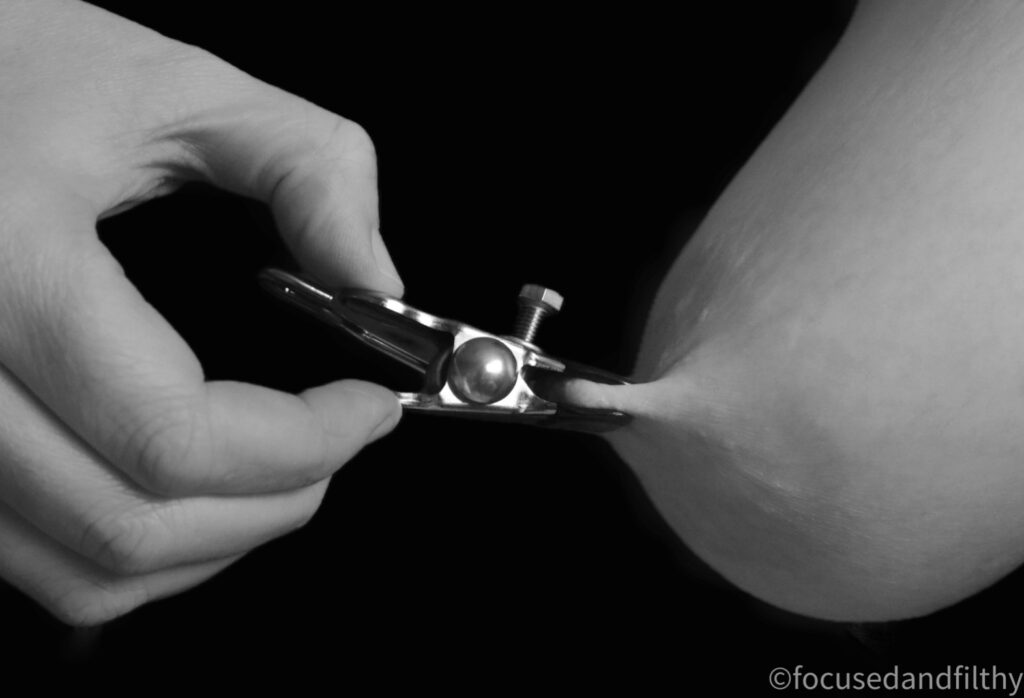 A close up black and white photograph of a hand pulling a metal nipple clamp that is attached to a female nipple and breast. The nipple is squeeze quite flat and the breast is being pulled forward.  