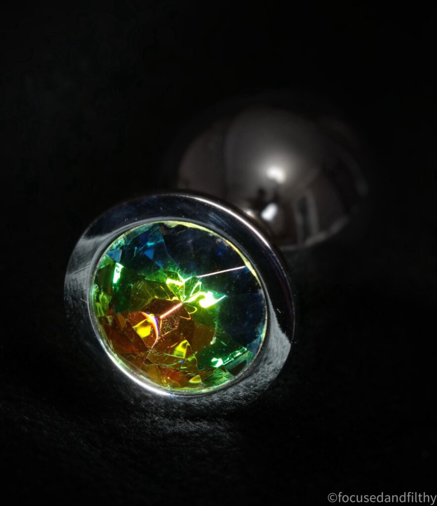 A close up photograph of the shiny jewellery end of a metal butt plug. The jewel is multicoloured and the background of this photo is completely black. 