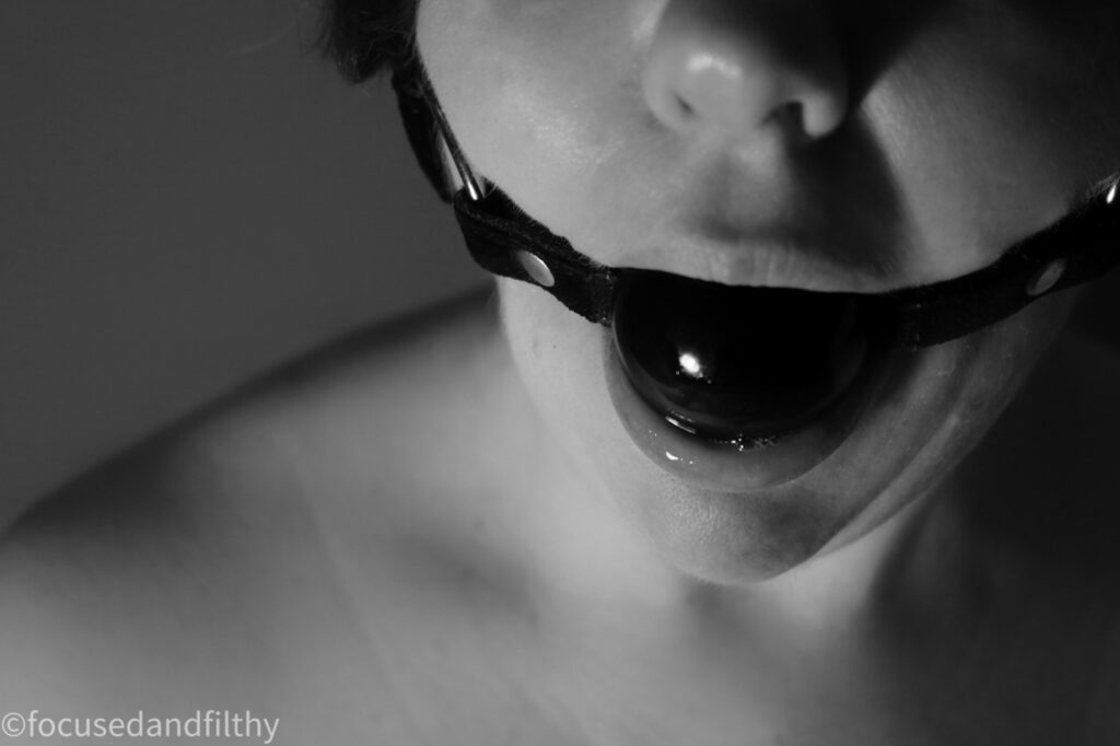 A black and white close up photograph of a woman’s mouth with a large black ball gag in. The saliva is just starting to glisten and pool around the ball. 