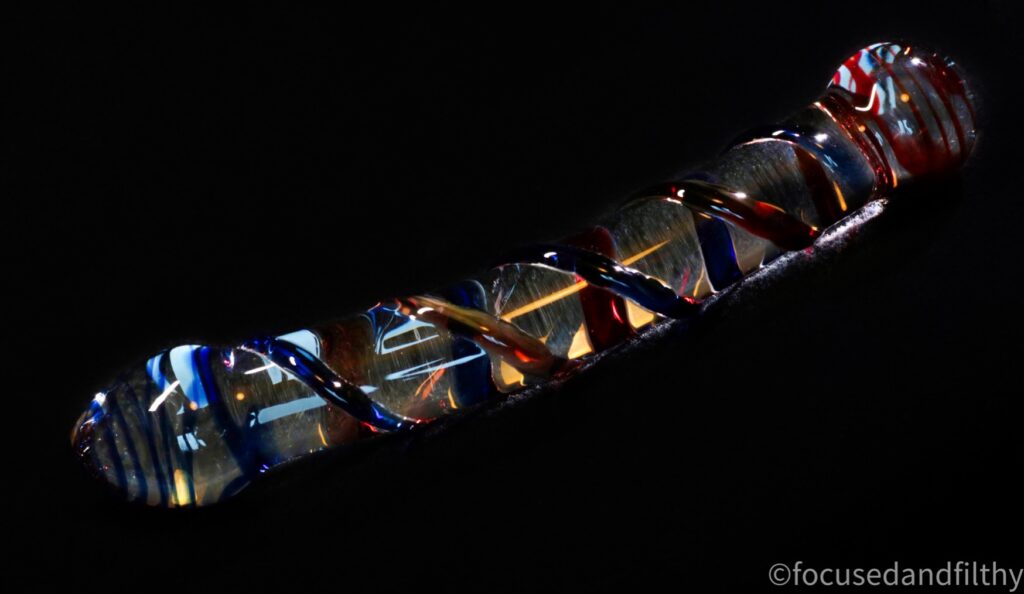A photograph of a glass doldo that has red and blue ribbon type ribbing running up the side with slightly bulbous edges the background is very dark.  