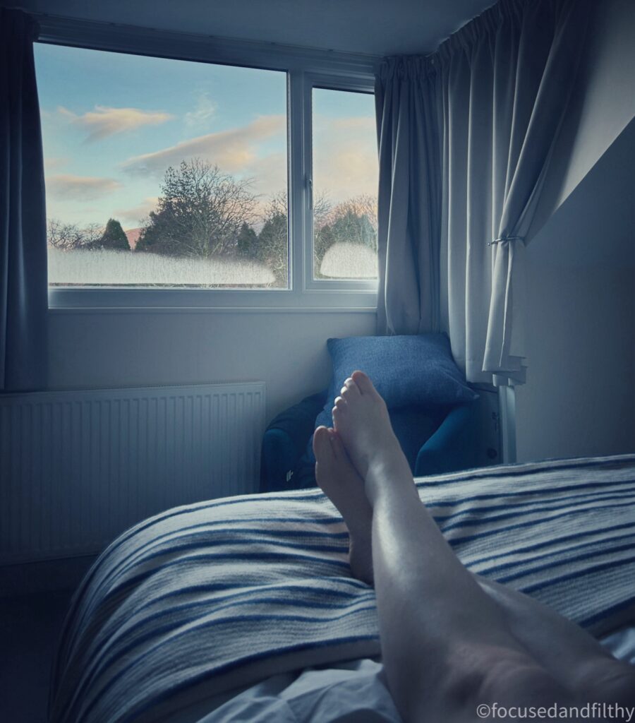 A POV colour photograph looking down my legs on a bed and out a window to see blue skies, trees and snow topped hills. 