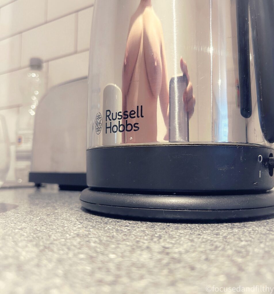 A close up colour photograph of a mirror reflective base of a silver Russell Hobbs kettle.  Reflected in the shiny surface is a female naked torso and she’s holding a mobile phone in one hand a white cup in another 