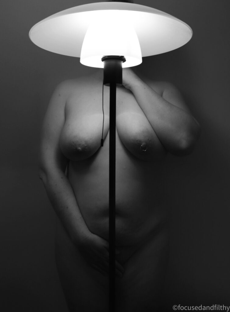 Black and white photograph of a standing lamp with a thin black metal pole and a domed top. Standing directly behind it is a naked woman. Her head is obscured by the domed light but her body is symmetrically placed behind the thin pole. She is curvy with a pierced left nipple, one handing is down covering her muffled and the other is up towards her face. 