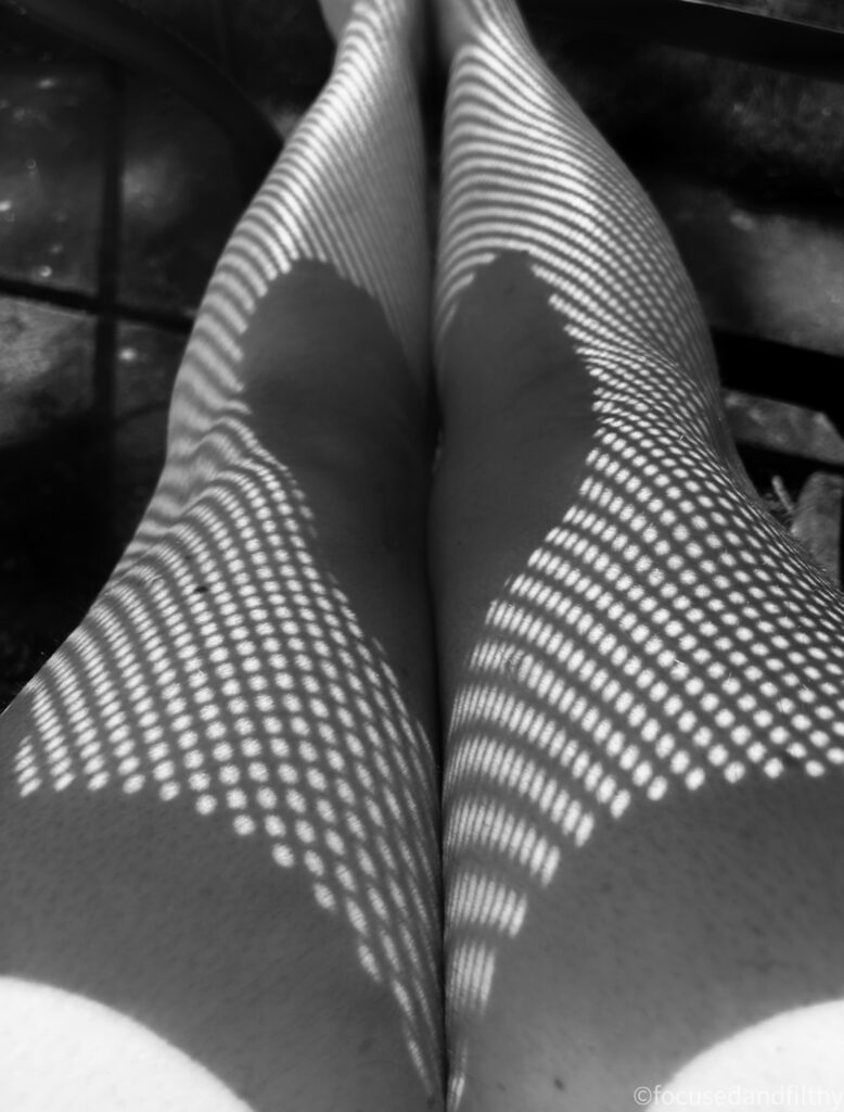 A black and white image of bare legs under a metal outdoor table. The table makes shadows that look like fishnets on the legs and there is a heart shaped shadow right across the middle. 