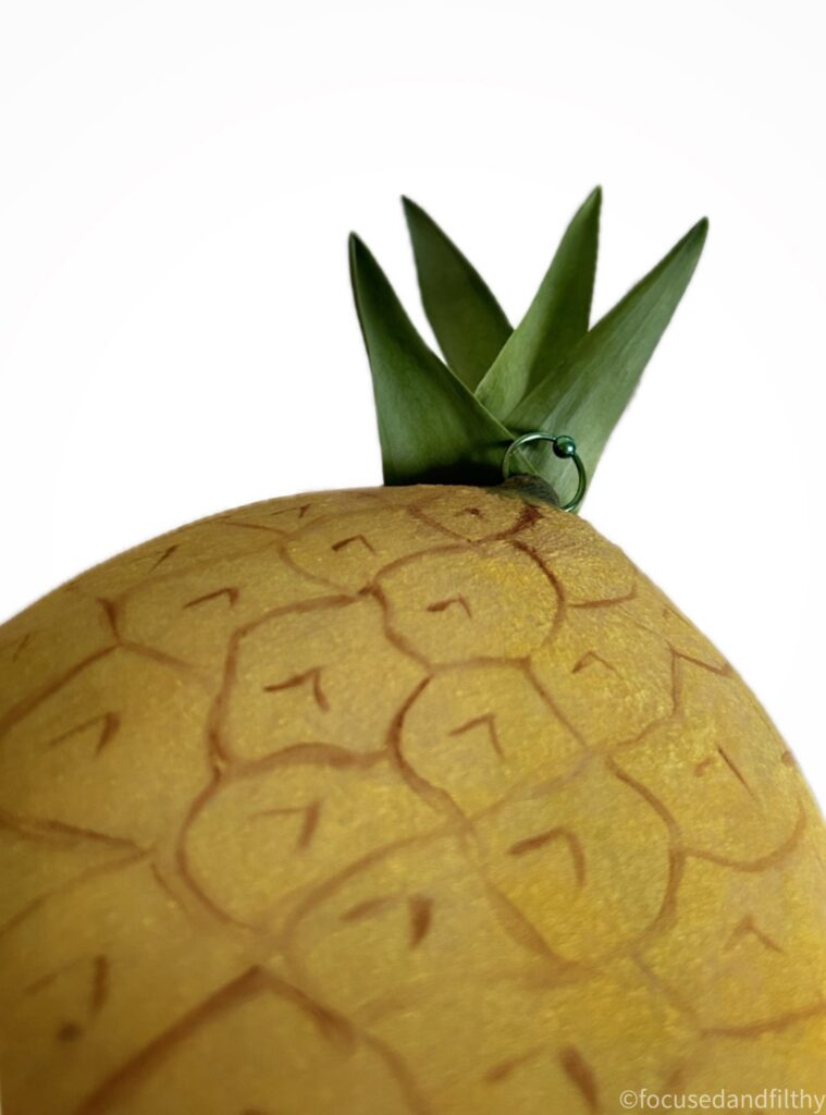 A close up photograph of what looks like a pineapple. But on closer inspection there is a green nipple ring among the spiked leaves at the top and the yellow and brown seems to be body paint on naked left breast. 