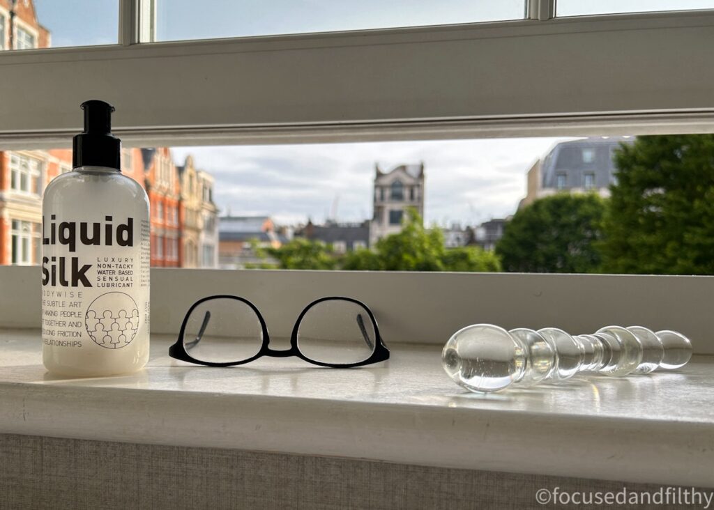 A photograph of a windowsill looking out onto a Soho square in London. On the windowsill is a bottle of nearly empty liquid Silk lube, a pair of men’s glasses with black frames and a glass dildo which is bumpy. 
