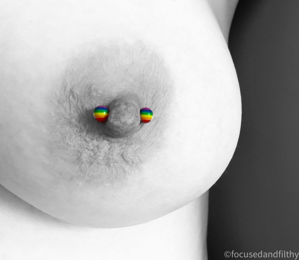 A black and white close up photograph of my large left breast apart from a colourful nipple bar that has rainbow coloured balls at each end. 