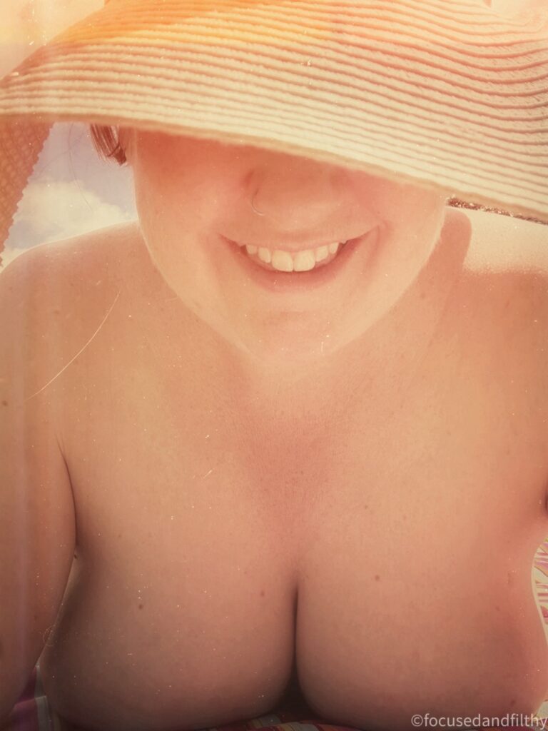 A colour photograph look at me laying naked on my front on the ground.  You can see the roundness of my breasts pressed against the ground and my smile but the rest of my face is covered by a large straw sun hat. 