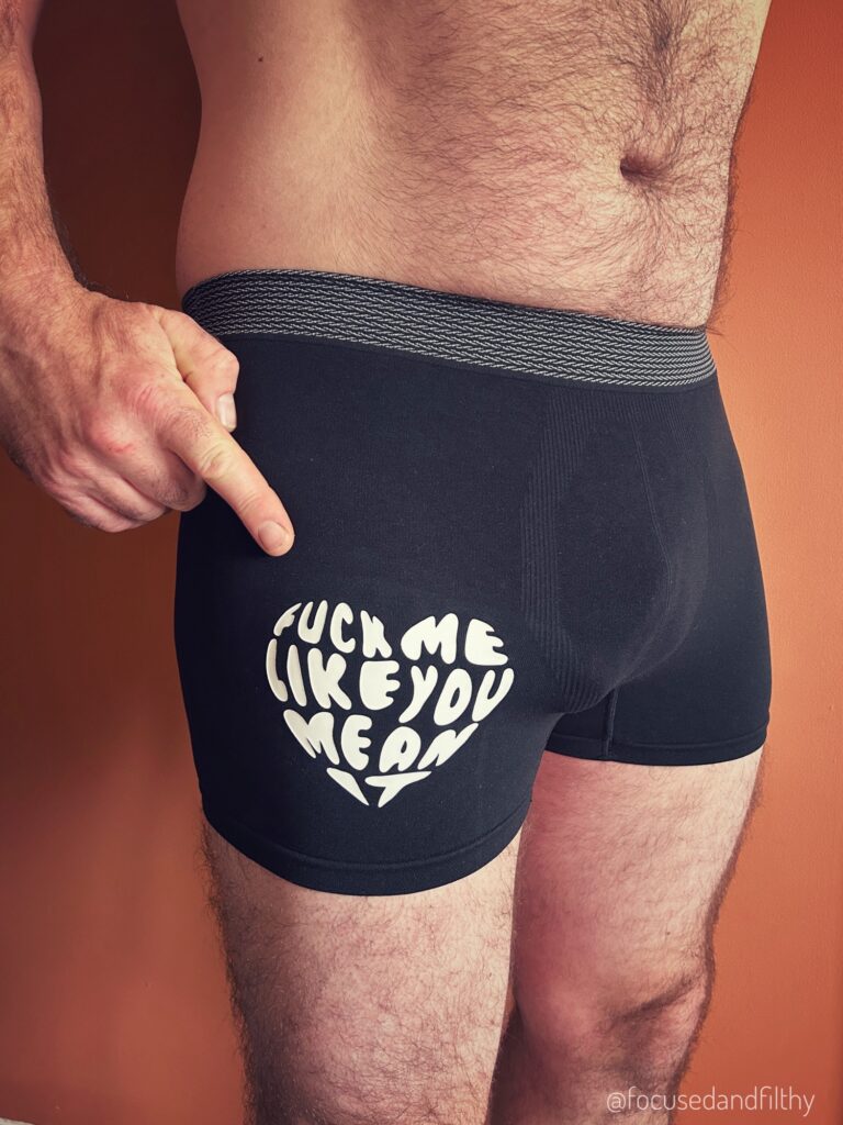 A colour photograph of the middle part of my man wearing just a pair of black jockey shorts. He is pointing to a large heart shape logo on them that says Fuck me like you mean it.  