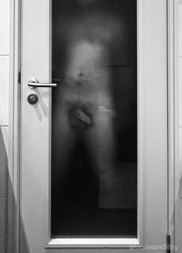 Black and white photograph of a bathroom door. The middle section has frosted glass in it and on the other side is dark but there is the middle section of a naked male body pressed up against it showing his cock. 