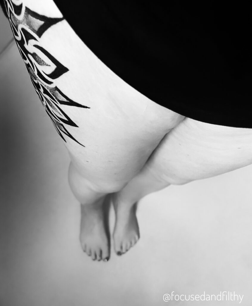 Black and white photograph looking down my body at my legs.  On my right thigh there is the pointed edge of a mandala style tattoo just creeping around onto the front of my thigh. Hinting at a larger piece.  