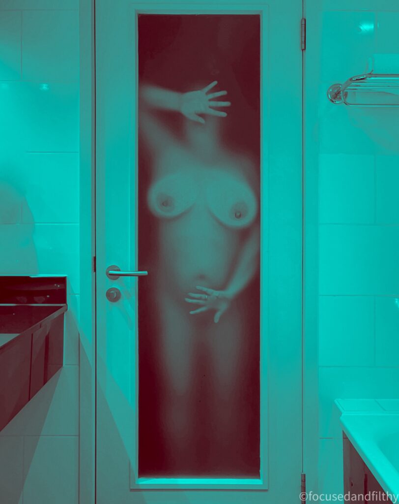 A photograph all edited into a turquoise monotone of a naked woman pressed against a frosted glass door so her hands are covering her face and muff, and her large boobs are pressed squished against the glass 