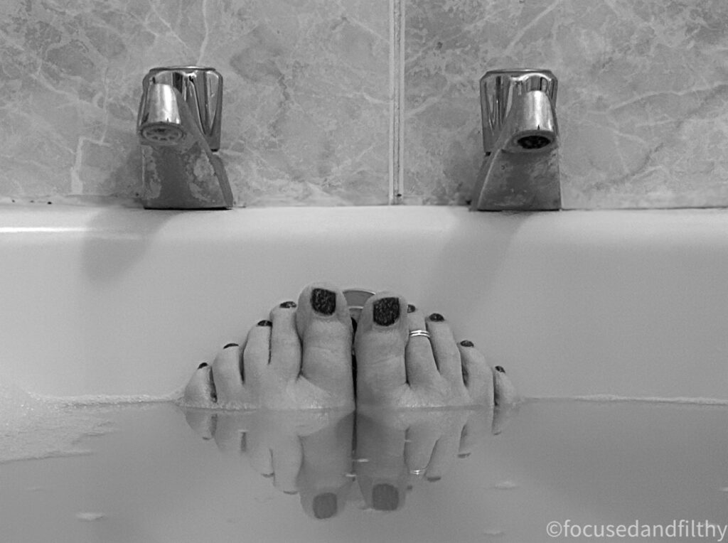 Black and white close up photograph of my toes against the bottom of the bath. You can just see the taps and my toes reflected back in the creamy murky water. I have darker nail varnish on my toes and a shiny toe ring on one toe. 