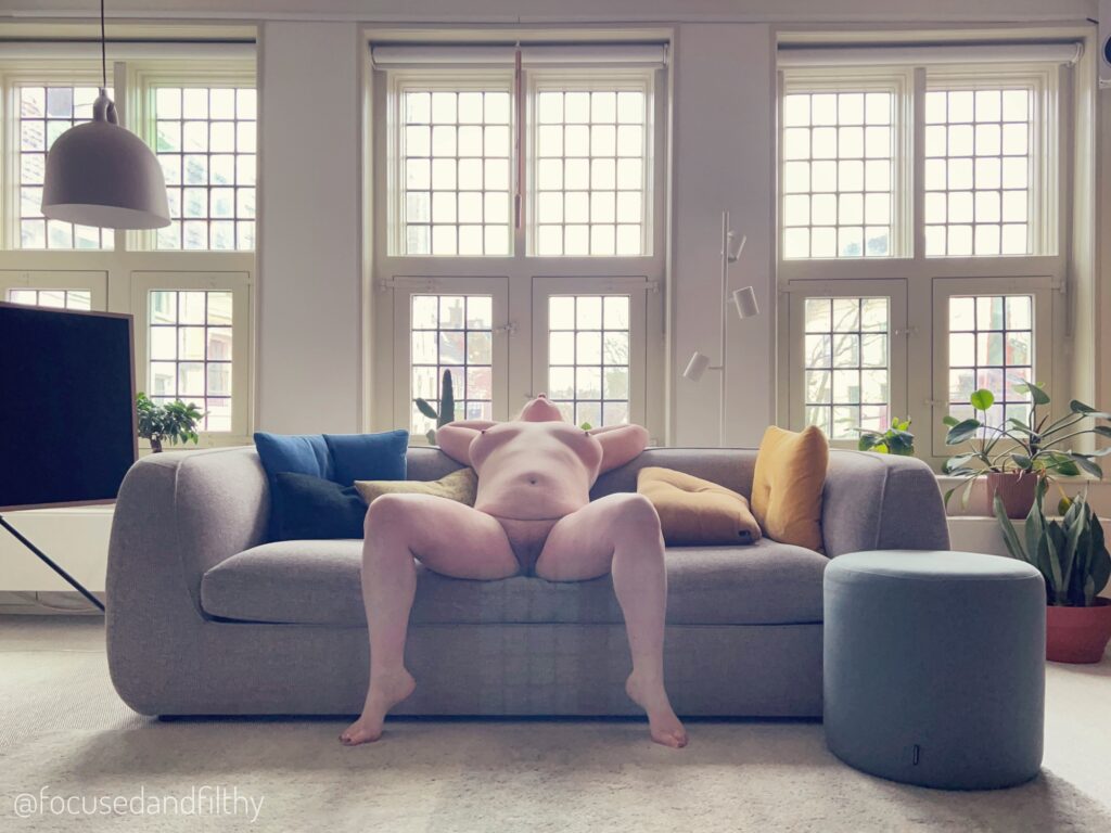 Image of me sitting naked on a large grey sofa with my legs wide and my head thrown back. The room is stylish but with old style leaded glass windows behind 