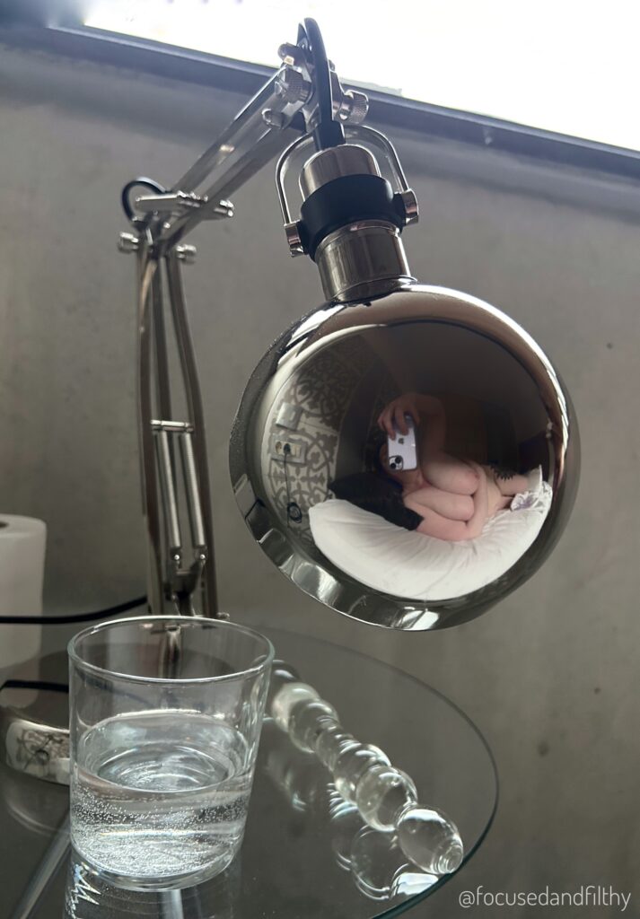 Photo of a shiny round bedside lamp and in the reflection is my naked torso and me holding my mobile phone to take the image.  On the bedside table there is also a glass of water a glass dildo. 