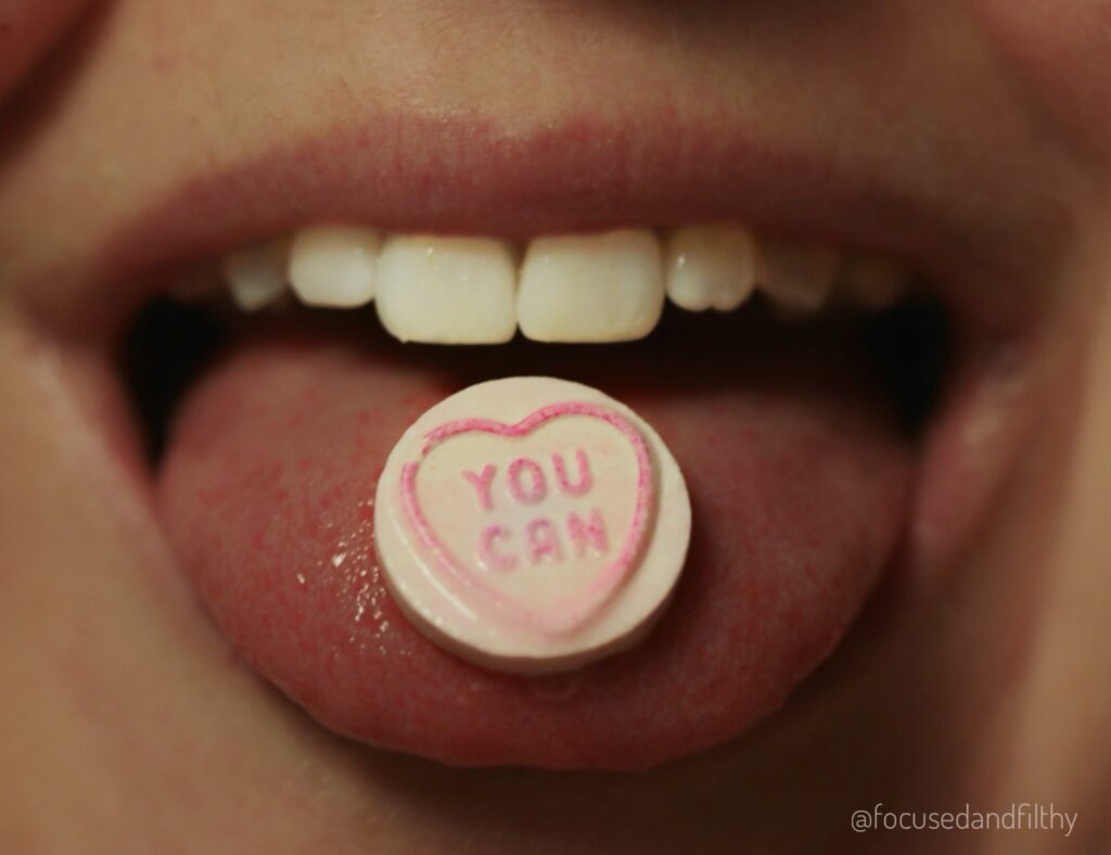 Close up photograph of my mouth and I’m sticking out my tongue and on my tongue is a love heart sweet that says ‘You Can’ on it. 