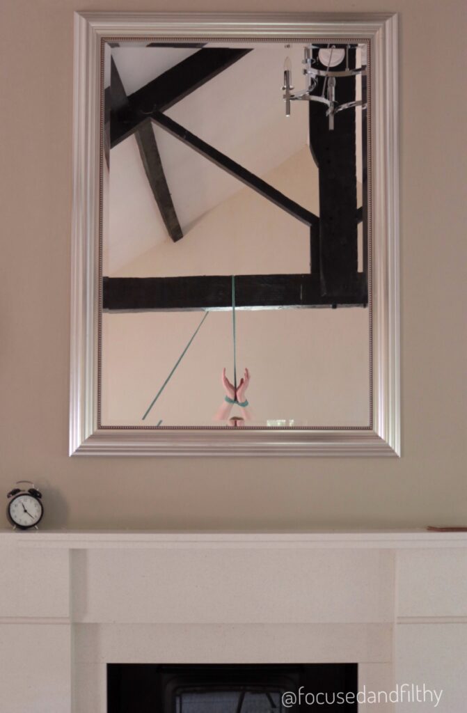 Photograph of a nice white fireplace with a large mirror above it. Reflected in the mirror is large dark ceiling beams and a rope hung from them attached to my hands held up high.