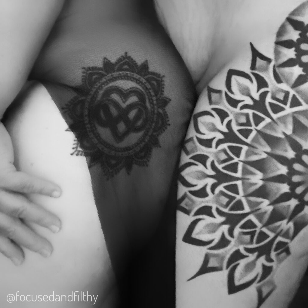 Black and white close up photo of two ladies right thighs, both have Mandela tattoos on. One is also got some sheer black floaty material over it the other skin is paler and a larger bolder spikey sun type Mandela tattoo one it.