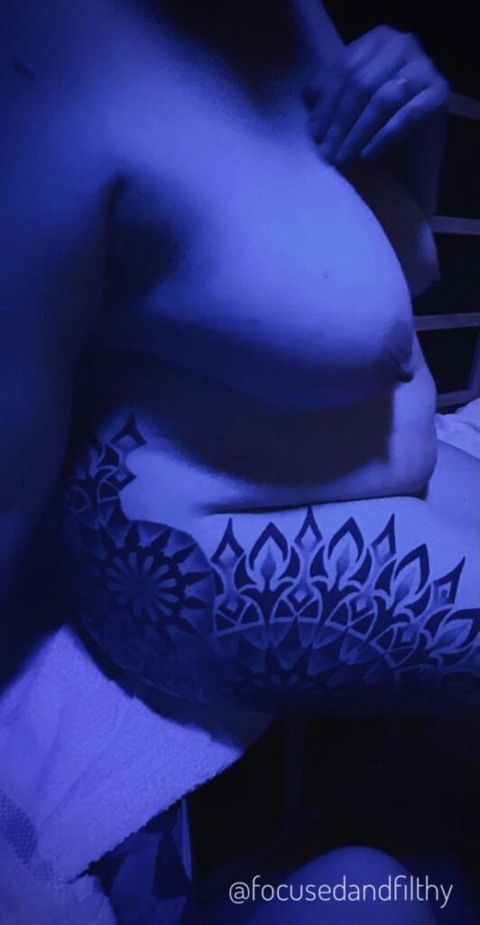 Photograph all the colour of blue of the side of me sitting in a small wooden sauna. You can see the tattoo on my hip and arse like a giant cogwheel contrasting against my pale skin