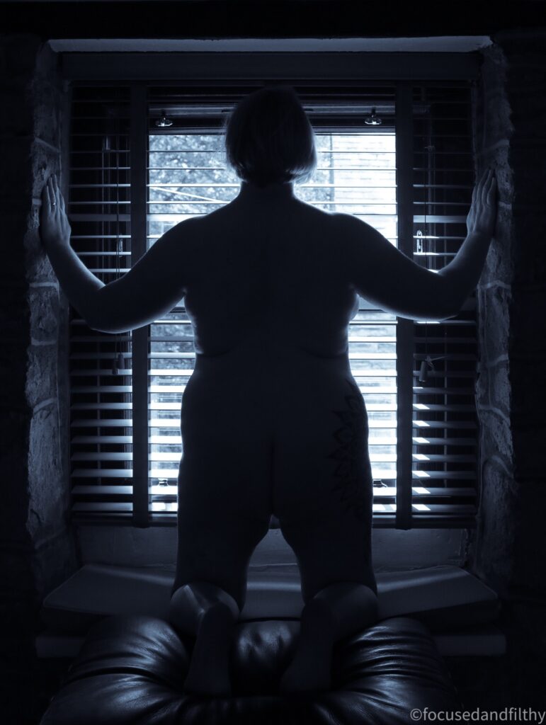 Black and white silhouette photo of me kneeling and looking out through a square window covered with wooden blinds.  