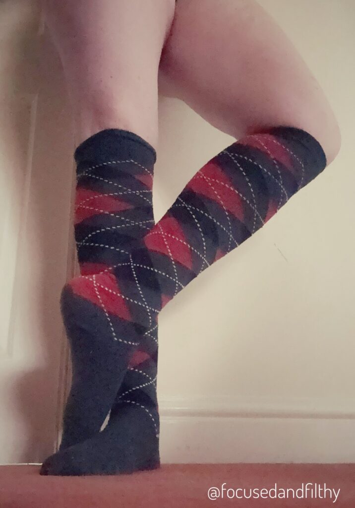 Photo of my legs (on knee out and that foot on its toes) wearing only knee length socks that are grey black and red in a golfer style diamond pattern. 