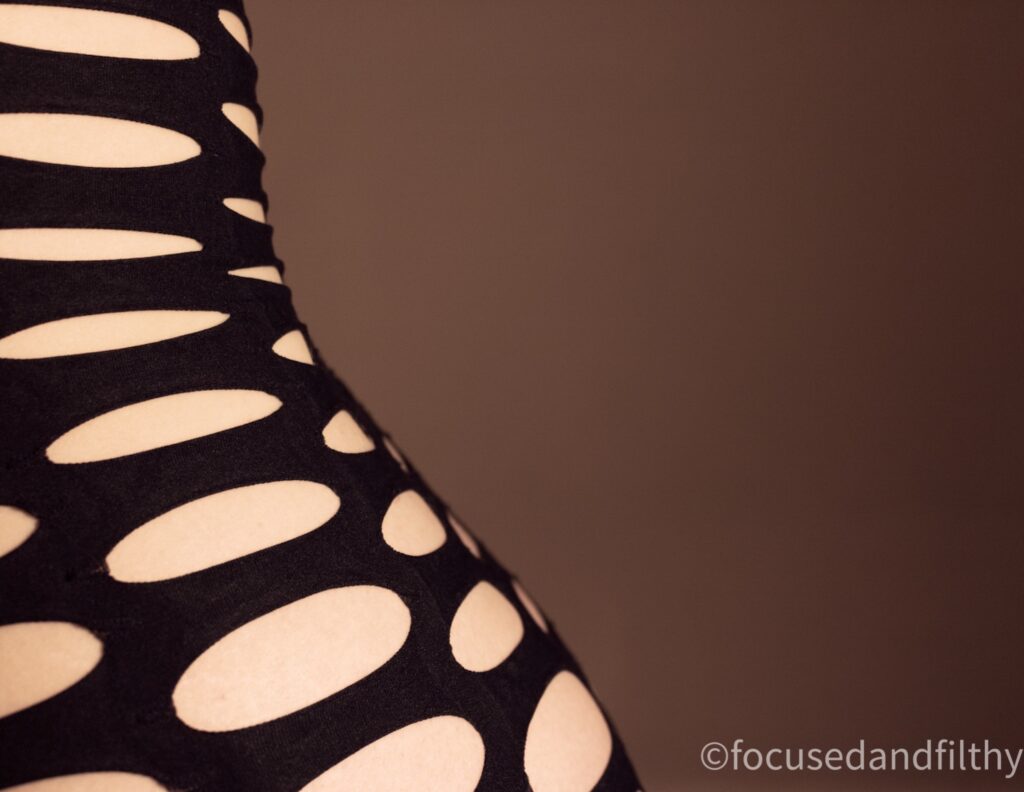 An abstract photo of the curve of my back wearing a bodystocking that has large oval shaped holes in it. The curve is on the left of the photo like a dropping curve of a graph. 