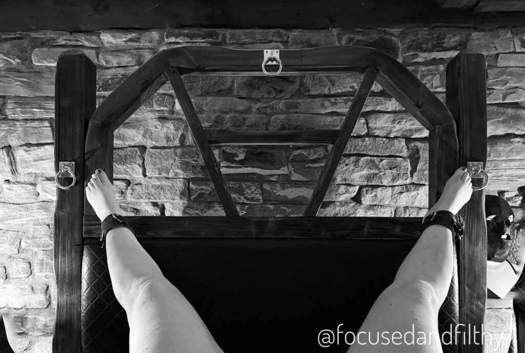 A black and white photo from my point of view, looking at my naked legs cuffed to the corners of a wooden headboard. 