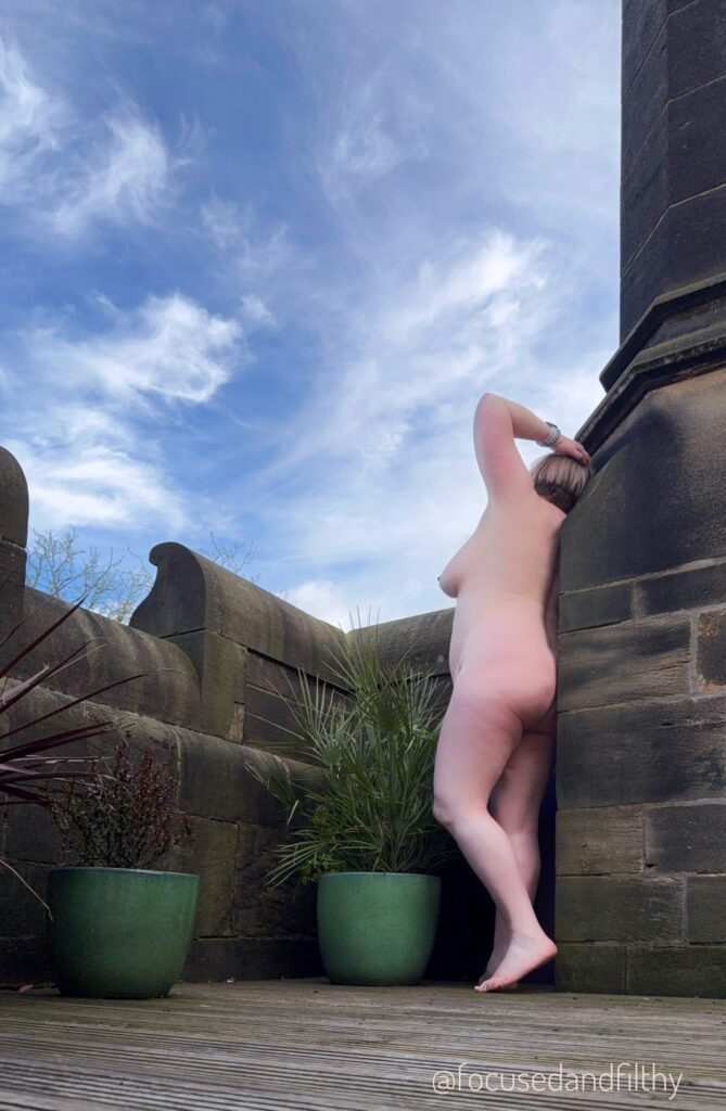 A photograph looking up from a wooden floor at a darkened stone wall of a roof top terrace with a naked me standing up against the stone. My arm is raised and my naked side just shows my left breast and curve of my hip down to my leg. The sky is blue and peppered with white wispy clouds