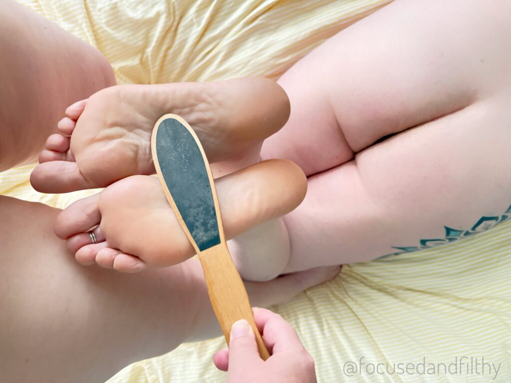 A photograph looking down onto a bed from her point of view. You can see me laying on my front my feet up and her standing above me holding a small wooden paddle to  my soles. 