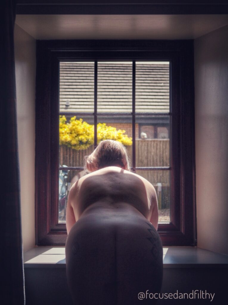 A photograph of me looking out a window.  I’m naked and you can see my arse sticking out. The window shows a gravelled area with cars in it and the sunshine is streaming in. 
