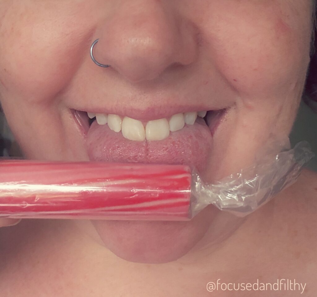 A close up photograph of my mouth where I am sticking out my tongue and I have a stick of bright pink rock (still in its cellophane) held against the tip 